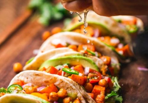 Tacos filled with chickpeas and vegetables are being garnished with lime juice on a wooden board with sliced limes beside them.