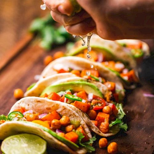 Tacos filled with chickpeas and vegetables are being garnished with lime juice on a wooden board with sliced limes beside them.