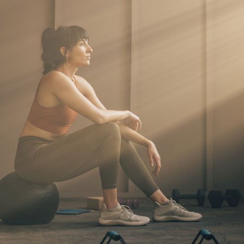 A person in workout attire sits on an exercise ball, with gym equipment nearby, bathed in soft sunlight streaming through a window.