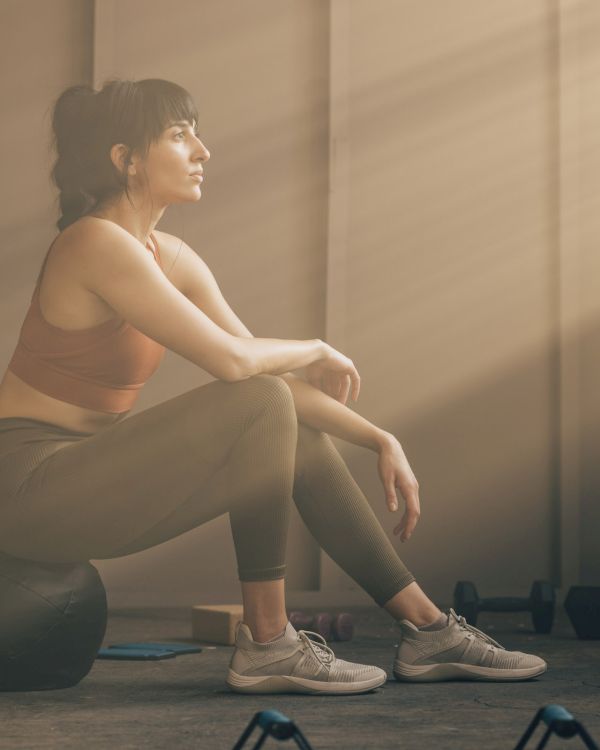 A person in workout attire sits on an exercise ball, with gym equipment nearby, bathed in soft sunlight streaming through a window.