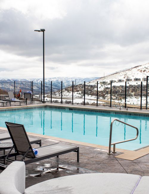An outdoor pool surrounded by lounge chairs, with a snowy mountain landscape in the background, beneath a cloudy sky.