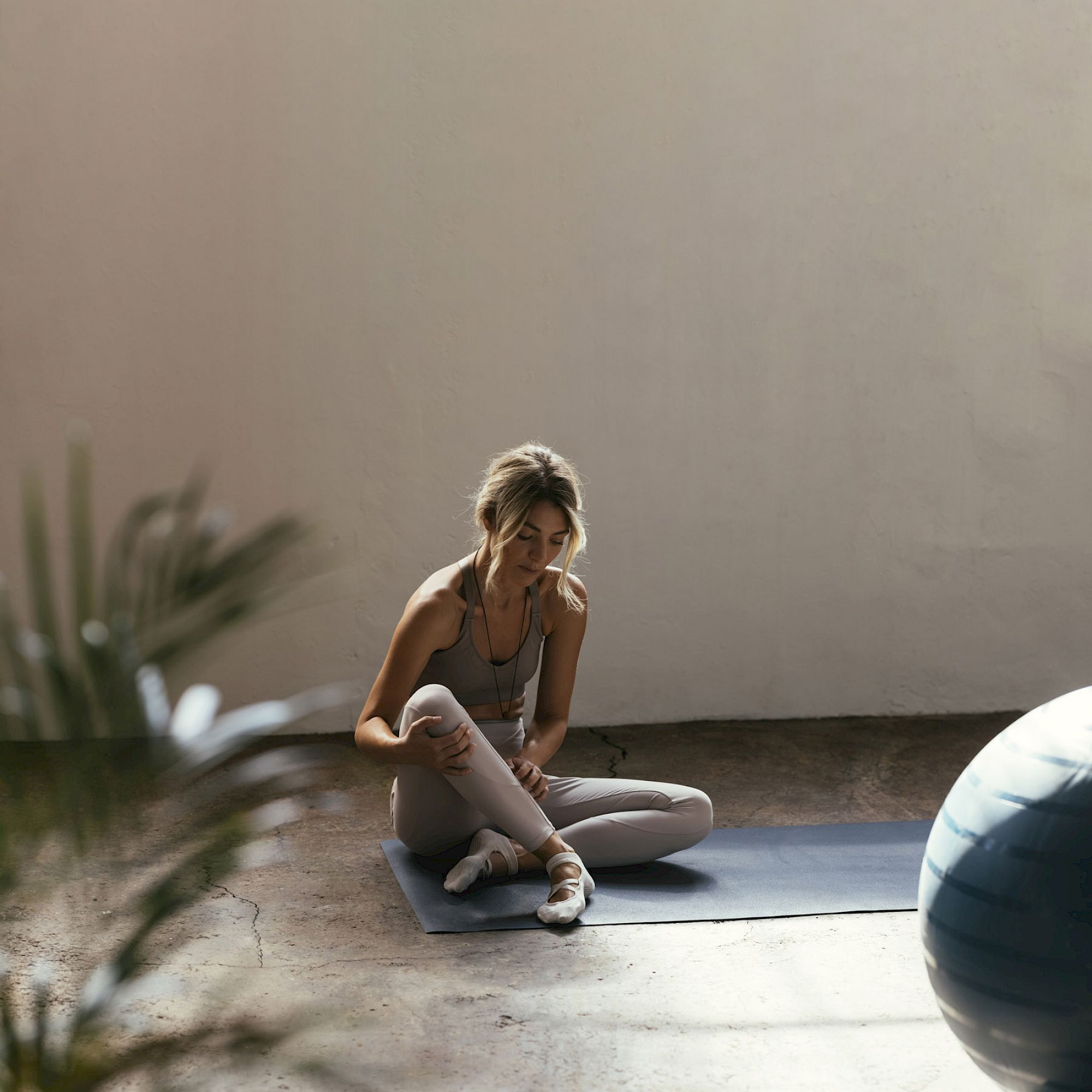 A person sitting on a yoga mat, stretching on the floor, with a large exercise ball nearby in a sunlit, serene space.