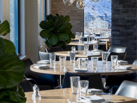 A stylish restaurant with neatly set tables, elegant lighting, large windows, plants, and a mountain view outside the window.