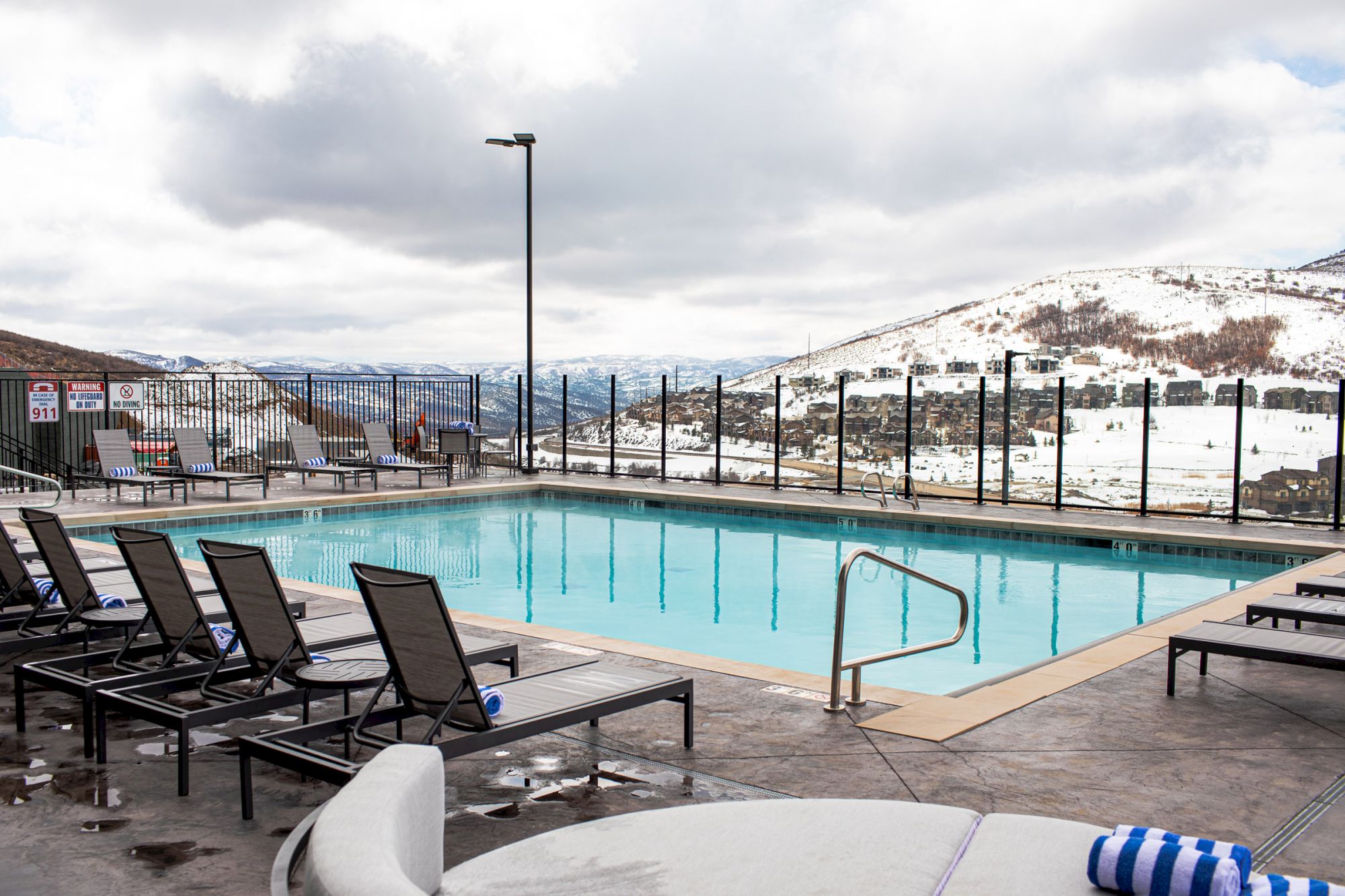 A poolside area with lounge chairs overlooks snowy mountains under a cloudy sky.