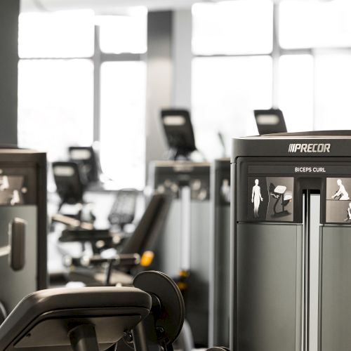 The image shows a gym with various exercise machines, including weight training equipment, in a well-lit room by large windows.