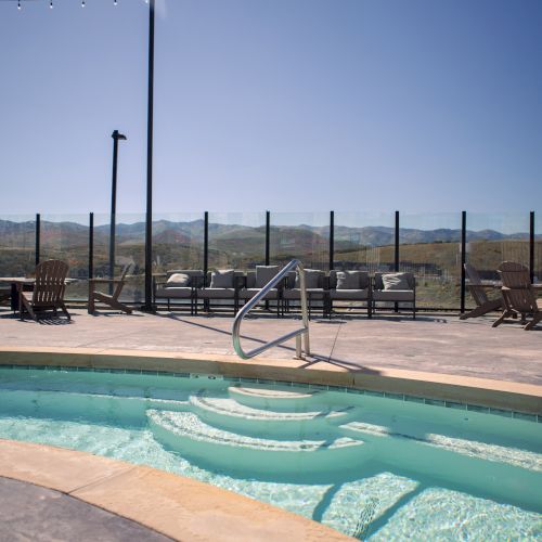 A small outdoor pool with a railing and surrounding lounge chairs, set against a backdrop of hills and a clear sky.