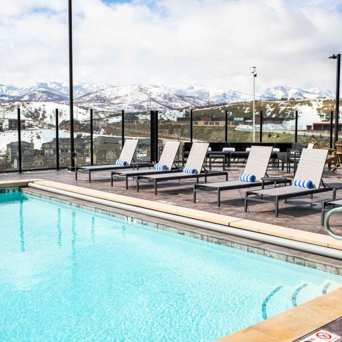 A swimming pool with lounge chairs sits under a cloudy sky, surrounded by glass fencing, with snow-covered mountains in the background.