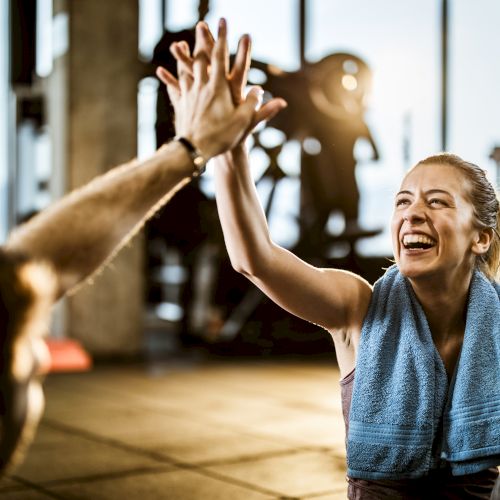 A person with a towel smiles and high-fives another in a gym, with exercise equipment visible in the background.