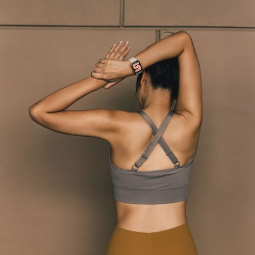 A person facing a wall, wearing a gray sports bra and mustard leggings, with arms raised over their head, and a smartwatch on the wrist.