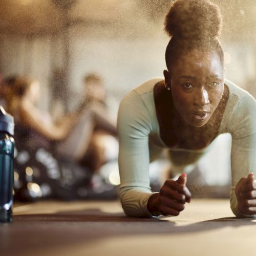 A person is doing a plank exercise in a gym, with a water bottle nearby. Others are seen exercising in the background.