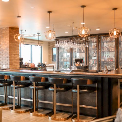 The image shows a modern bar with high chairs, glassware, warm lighting, and brick accents, creating a cozy and inviting atmosphere.