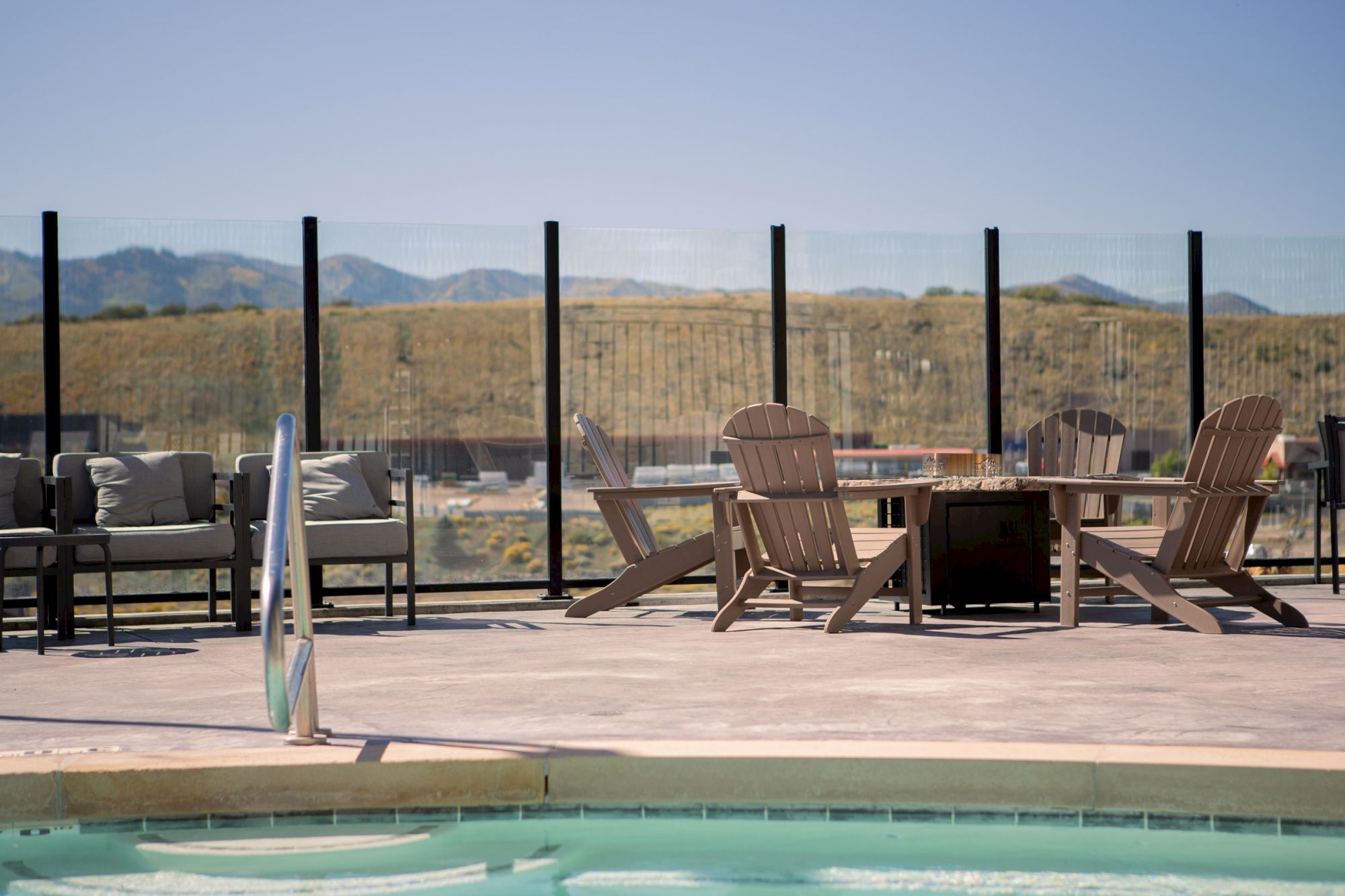 A poolside area with wooden chairs, a table, and mountains in the background under a clear sky.