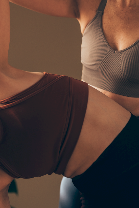 Two people practicing yoga; one assisting the other with a pose. They are wearing sports bras and leggings in a softly lit room.