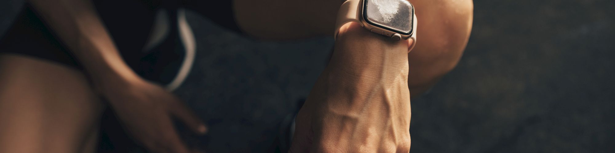A person wearing athletic shorts and sneakers sits, focusing on a smartwatch on their wrist, with a dark outdoor surface nearby.